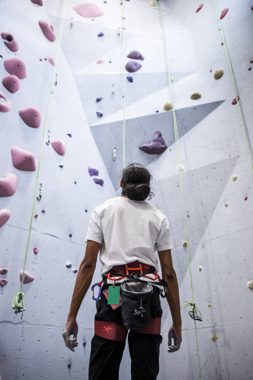 A climber in a white T-shirt and harness stands with their back to the camera, looking up at a tall grey wall of purple and pink holds