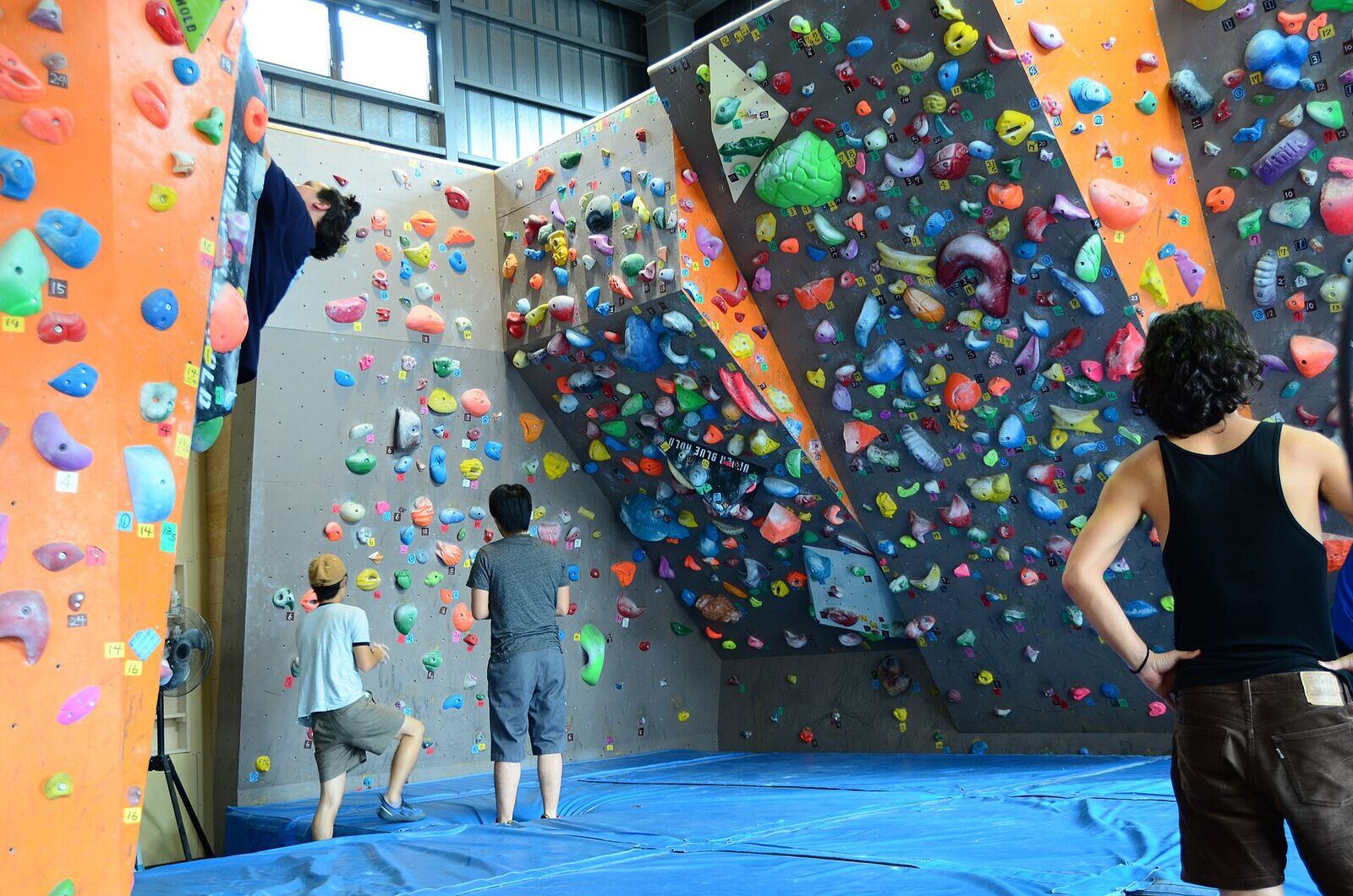 Friends stand on blue mats watching each other on a bouldering wall packed with bright holds, one climber reaching over the top of an orange pillar