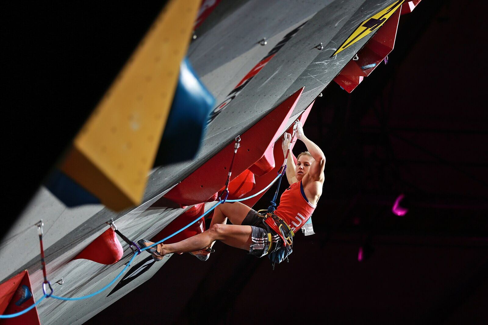 A competitor in a red vest hangs below a steep competition roof, rope clipped, against a dark arena