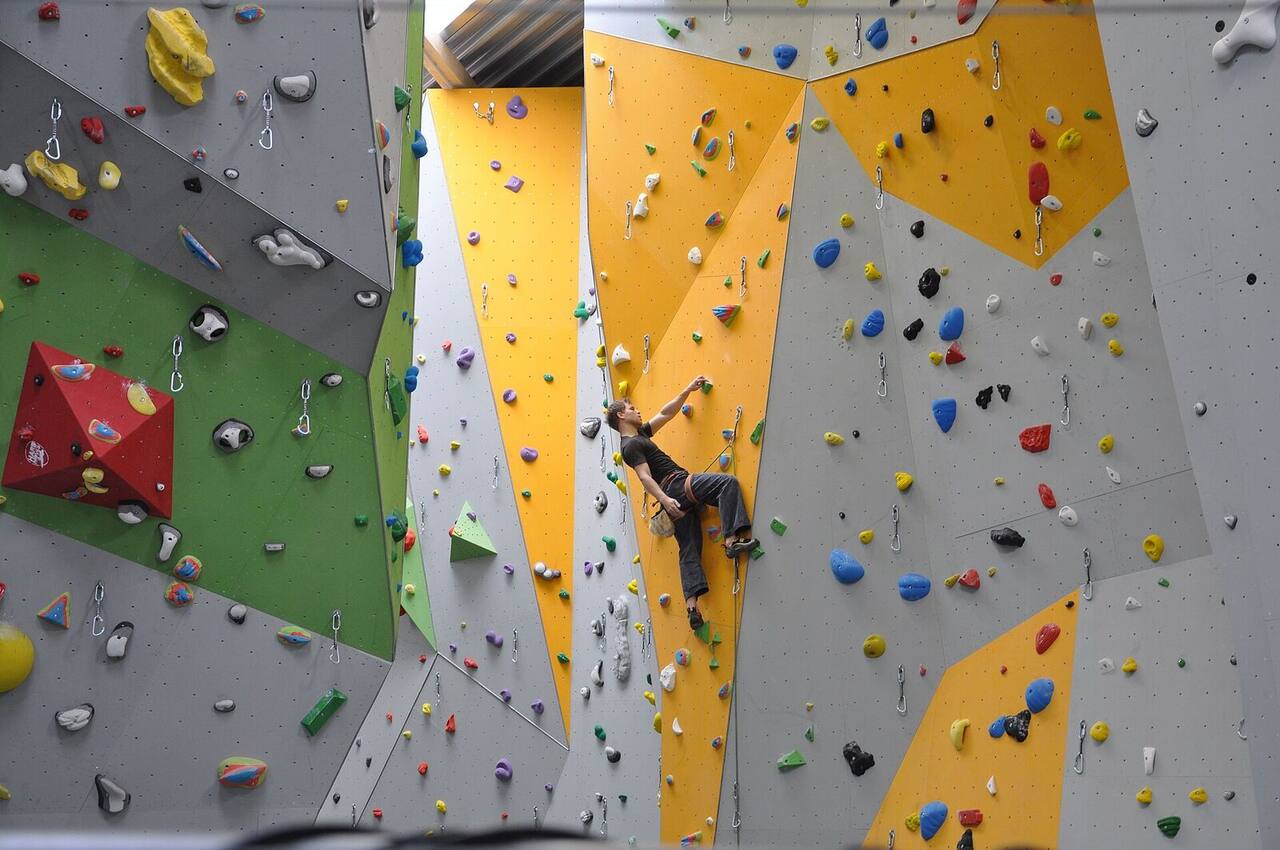 A climber leads up a yellow and grey wall, reaching for the next hold with the rope clipped below