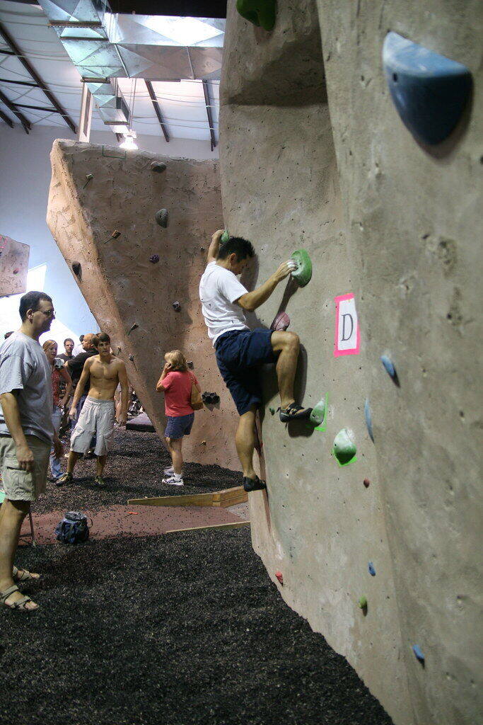 A climber works a beige bouldering wall at a local competition while others wait and watch